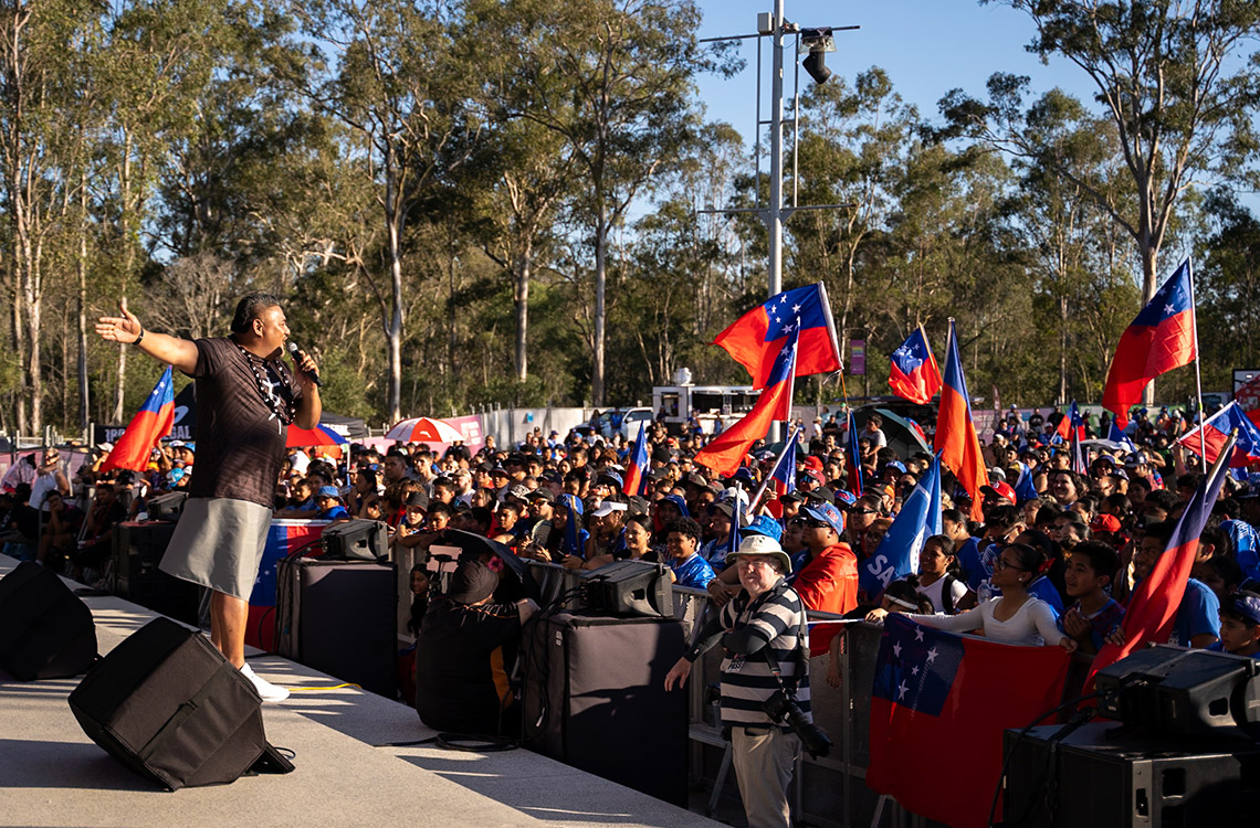 Samoan man on stage talking to a large crowd of rugby fans waving flags